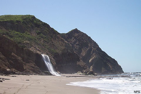 A waterfall cascades down a short cliff on left onto a sandy beach as waves wash in from the right. Two hills rise in the background.