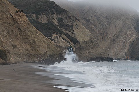 Pacific Ocean waves wash across a sandy beach from the right and crash against the base of a bluff at the center of the image. Just beyond the wave splash, a waterfall cascades on to the beach.