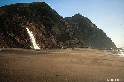 Water cascades down a short cliff from the left onto a wide sandy beach. Waves break on the right.
