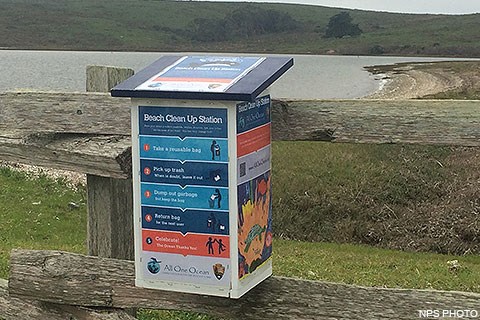 A colorful vertically-aligned box attached to a wooden fence with a beach in the background.