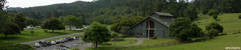 A gray barn-like visitor center surrounded by green fields and trees with a parking lot on the left.