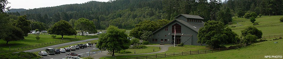 A gray barn-like visitor center surrounded by green fields and trees with a parking lot on the left. A gray barn-like visitor center surrounded by green fields and trees with a parking lot on the left.