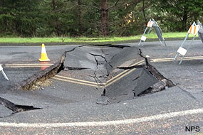A six-foot-wide, 14-foot-long depression filled with broken, collapsed concrete spans a two-lane road.