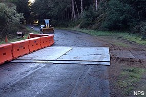 Concrete plates span the width of a two-lane road with orange K-rails on the left and a yellow front-end loader in the distance.