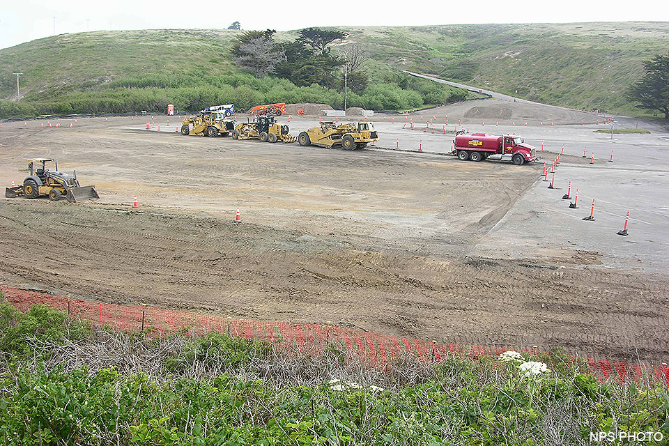 A front end loader, a road grader, an earthmover, and other heavy equipment are parked in a parking lot that is under reconstruction.