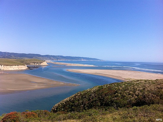 A photo looking down from a blufftop at an estuary winding between sand bars as it heads for an ocean bay in the distance.