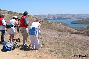 Four people, two of whom are wearing red vests, look toward a body of water in the distance through spotting scopes.
