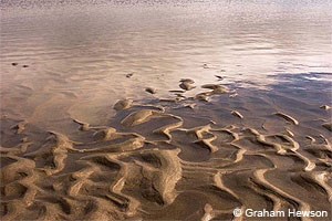 Ripples of sand at the tide's edge with clouds reflected in the water.
