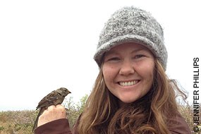 A woman with long blonde hair wearing a gray knit cap smiles at the camera while she holds a sparrow in her hand.