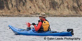 A kayaker wearing a red jacket and a yellow PFD paddles a blue, plastic kayak.