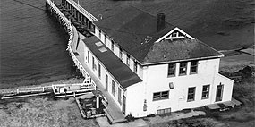 A historic black and white photograph looking down at a two-story-tall lifeboat station on the edge of a body of water.