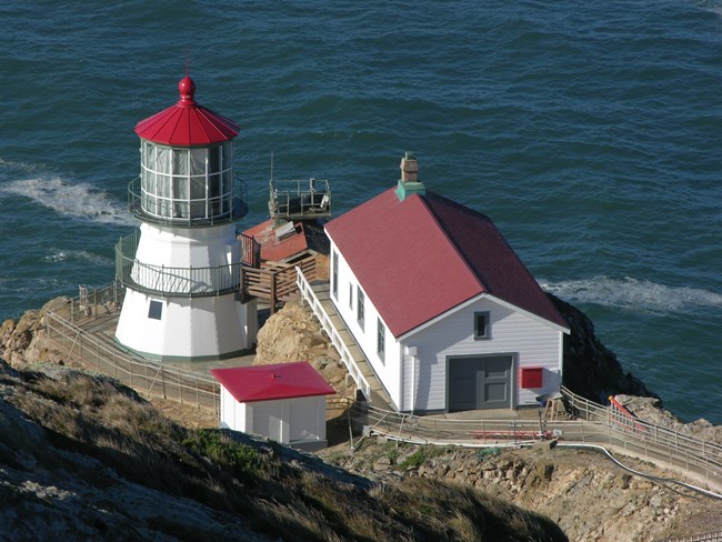 Four white-sided, red-roofed light station buildings sit on the edge of an ocean-side bluff.
