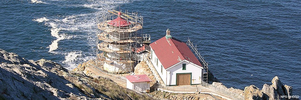 hree white-sided, red-roofed buildings perched on a rocky headland with the ocean in the background. The lighthouse tower is surrounded by scaffolding. hree white-sided, red-roofed buildings perched on a rocky headland with the ocean in the background. The lighthouse tower is surrounded by scaffolding.