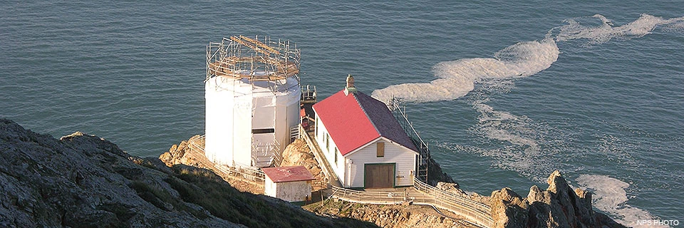 The Point Reyes Lighthouse surrounded by a nylon shroud on December 8, 2018. Three structures—a tower, a large rectangular building, and a small square structure—sit on a rocky headland above the Pacific Ocean. The roof and rafters of the lighthouse tower have been removed and scaffolding and white fabric surrounds the tower.