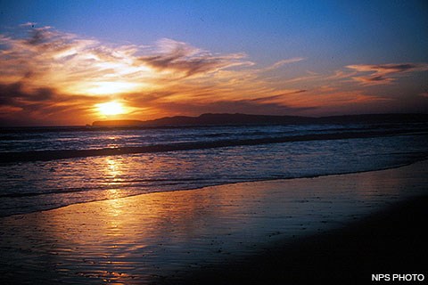 Light from the setting sun filters through clouds over headlands in the distance and waves washing onto a beach in the foreground.