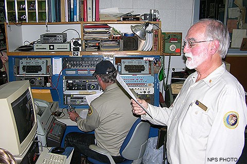 A seated volunteer surrounded by old computers and electronic equipment uses a Morse key to broadcast a Morse code radio message while a volunteer standing on the right reads the message aloud to attendees.