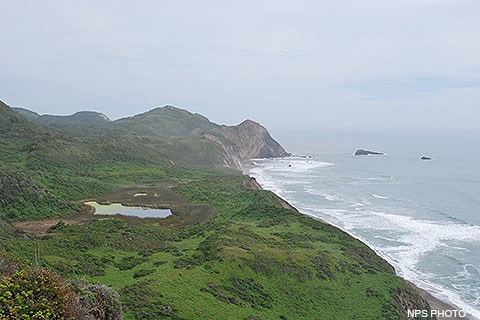 Below overcast skies, Pacific Ocean waves wash in from the right, breaking against a rocky headland in the image's center. Vegetated hills and valleys fill the lower left quadrant of the image, with a small lake filling a depression in the center right.