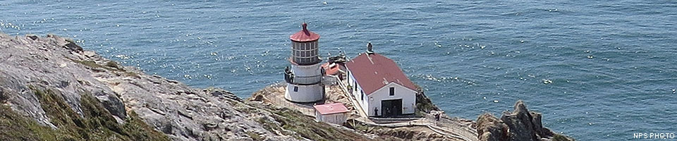 The Point Reyes Lighthouse on April 9, 2017. On the edge of the Pacific Ocean, a white, three-story tower with a red roof stands adjacent to three other red-roofed, white-sided buildings on. Barren rocks slope down to the lighthouse on the left.