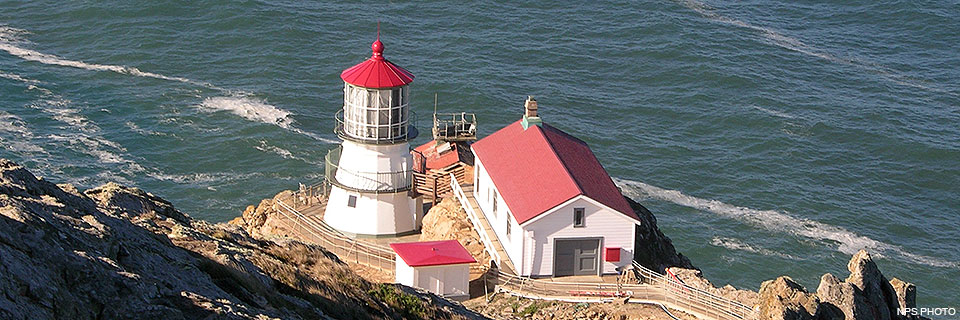 The Point Reyes Lighthouse on October 21, 2019, shortly after contractors had removed a nylon shroud used during a restoration project. Four red-roofed, white-sided buildings sit on a rocky headland surrounded by water.