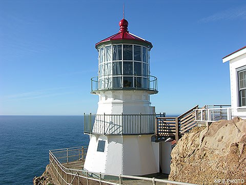 A three-story-tall, white-sided, red-roofed lighthouse perched on a rocky headland with the ocean in the background.