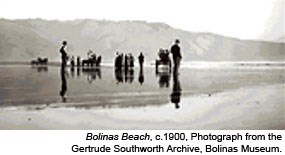 A black-and-white historic photograph of over a dozen people standing on a beach with a ridge rising in the background.