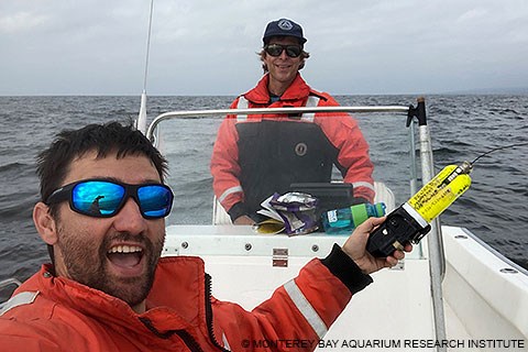 A man wearing an orange coat and sunglasses holds a yellow and black-colored cylindrical device while on a small boat while the driver of the boat stands in the background.