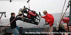 Two men on a boat maneuver a small robotic submersible hanging from a crane.