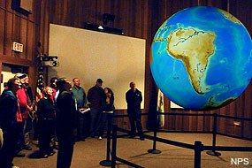 Inside a moderately sized room, a dozen people with a ranger look at a large globe of Earth suspended from the ceiling.