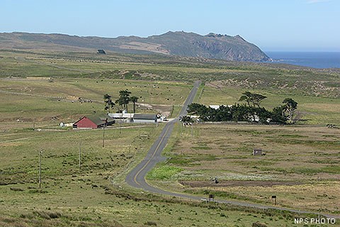 A narrow road winds through tan and green pastureland, through a cluster of buildings, and off toward a rocky headland on the edge of the ocean.