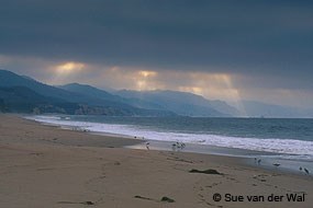 Sun streams through a few breaks in an overcast sky above a sandy beach in the foreground and haze-shrouded hills in the background.