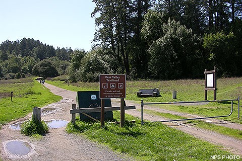 A wide foot path passes by trailhead signs and a metal gate. The trail passes through a meadow before entering a forest in the distance.