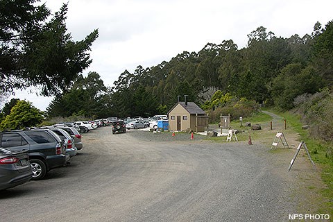 Many vehicles parked in a gravel parking lot on the left. A vault toilet building is located just right of center. A foot path heads off into the woods on the right.