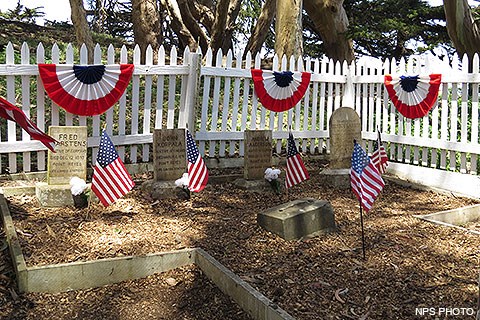 Five graves with off-white headstones surrounded by a white picket fence on which red-white-and-blue bunting is hung.