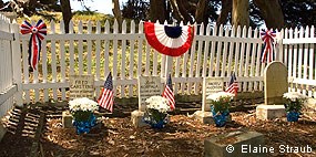 Four small white headstones in a small cemetery surrounded by a white picket fence on which some bunting is hung.
