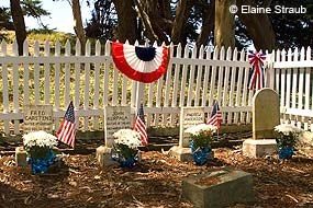Four small white headstones in a small cemetery surrounded by a white picket fence on which some bunting is hung.