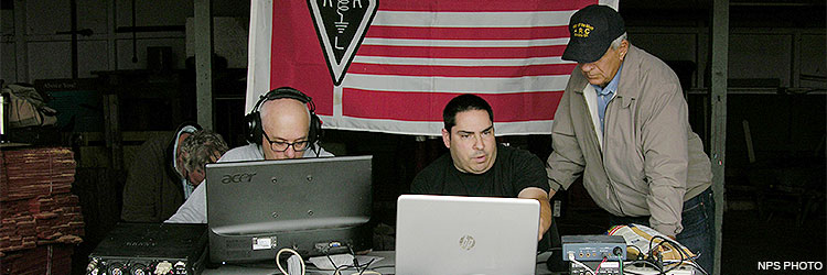 Three men sitting at a folding table on which there are laptops and radio equipment. A red, white, and black flag hangs in the background. Three men sitting at a folding table on which there are laptops and radio equipment. A red, white, and black flag hangs in the background.