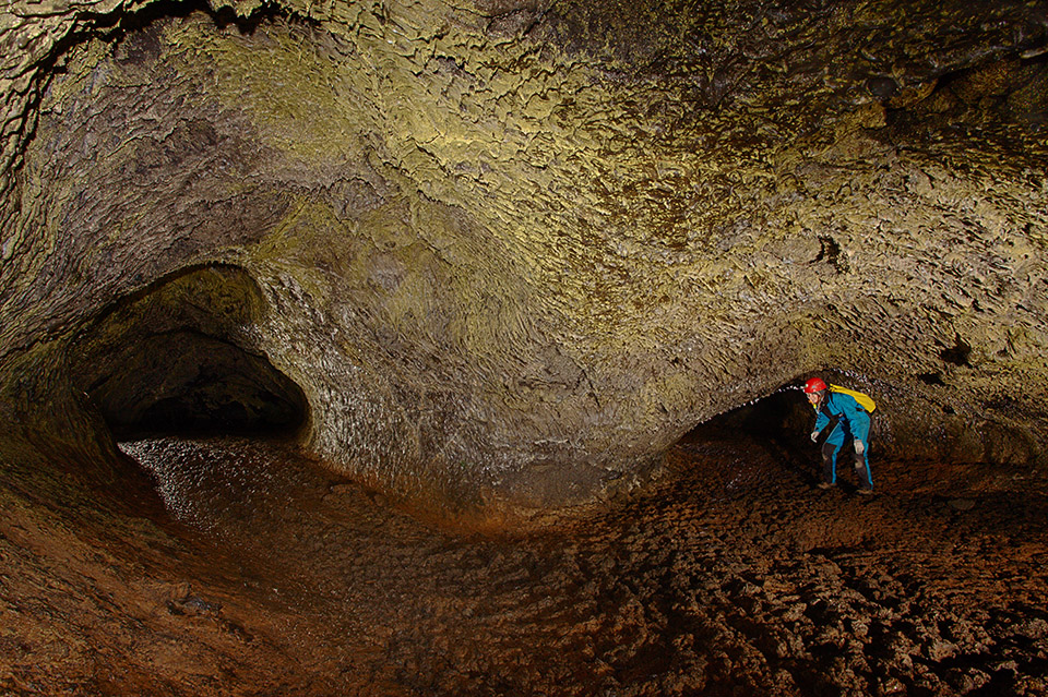 Rock wall inside a cave is mottled grey and has a yellow-tinged section in the middle. On the left and on the right are openings to additional caves. An individual dressed in a blue body suit with black patches from knee to toe, wearing a red hard hat, grey gloves and a yellow backpack crouches at the low entrance to the cave on the right. The foreground extending into the two cave entries is rust colored and appears mud-like with irregular clumps.