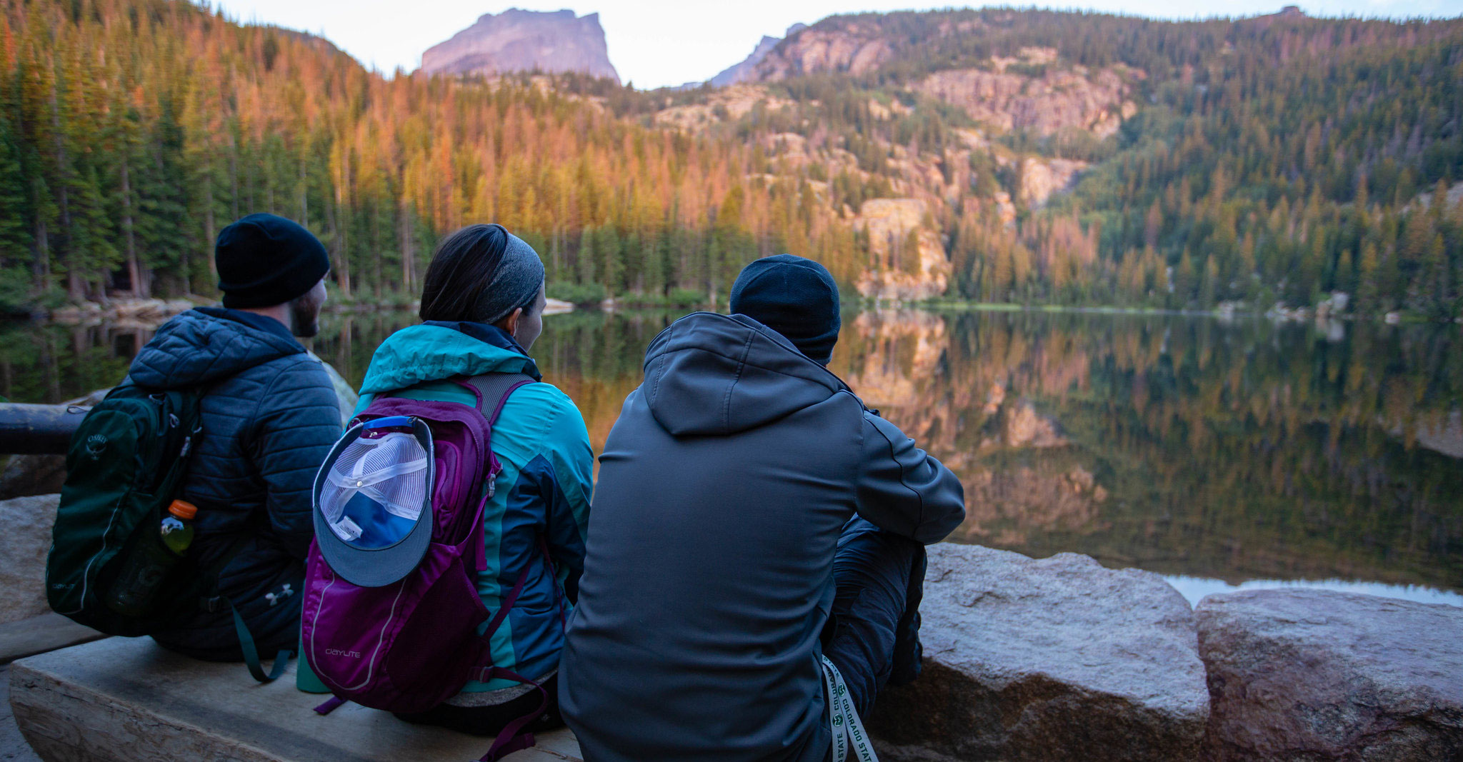 three people dressed in warm clothes sit on a wooden bench overlooking a lake a sunlit trees