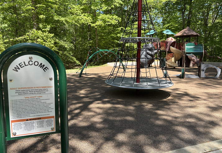 Children's playground structures and a welcome sign are shaded by a green forest