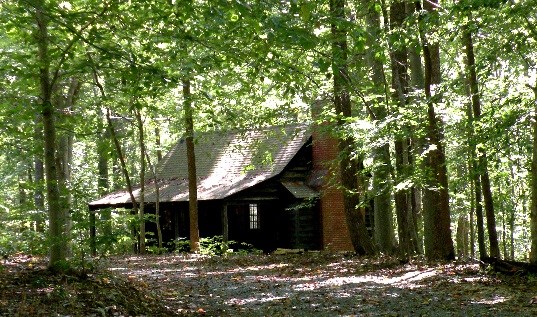 Wooden cabin with a porch and brick chimney shaded by green trees beside a gravel road
