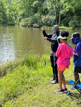 Family of 2 kids and an adult reeling in a fish on the grassy side of a lake in the forest