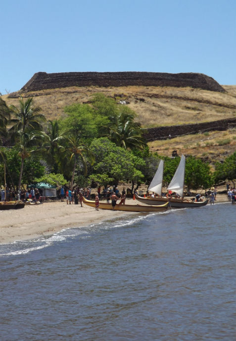 Visitors gather along Pelekane Beach during a Hawaiian Cultural Festival. Canoes along the shore with Pu'ukohola Heiau rising in the background.