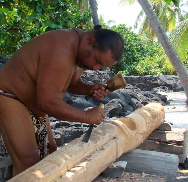 Man in traditional clothing carves a kiʻi image