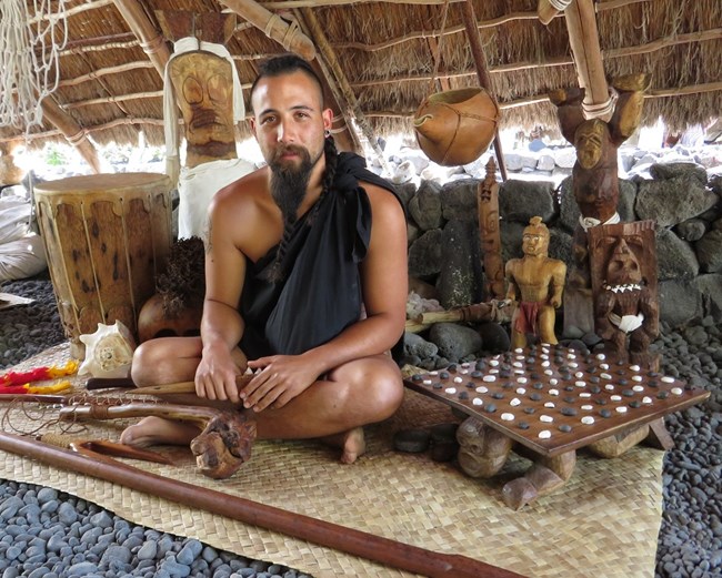 Man in traditional clothing sits among Hawaiian cultural items