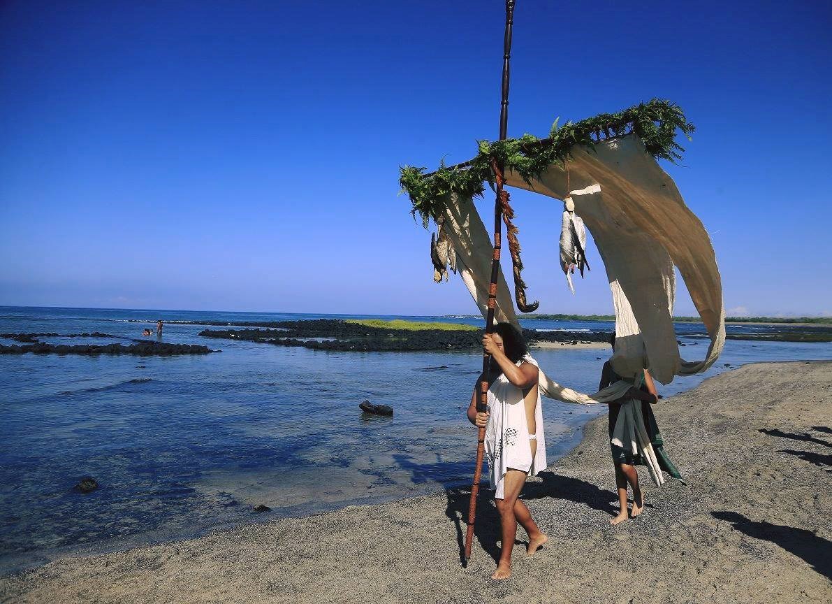 A man in traditional clothing walks the shoreline carrying the akua loa draped with white kapa cloth and a lei made of ferns adorns the crosspiece