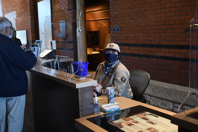 A volunteer in uniform smiles towards the camera as she interacts with visitors at a greeter desk.