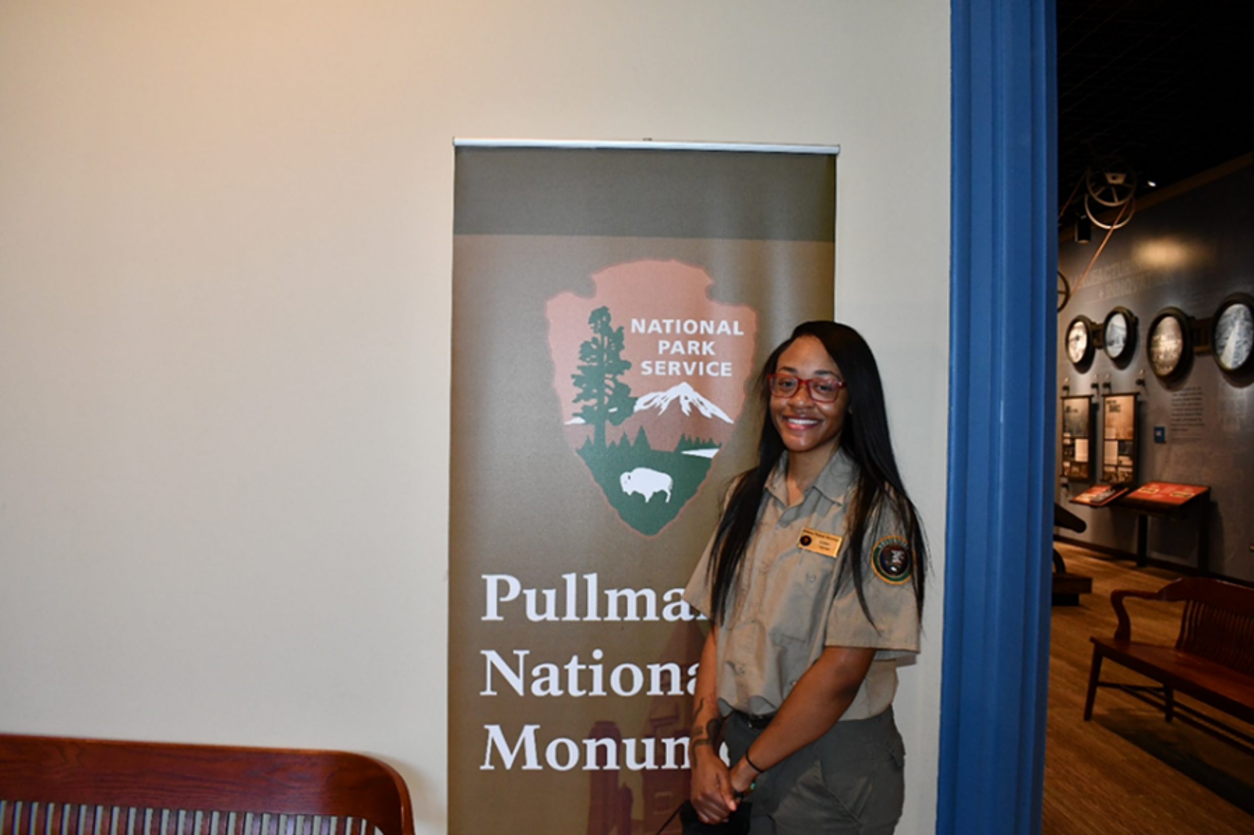 A volunteer in uniform smiles at the camera standing in front of banner with the NPS arrowhead and words "Pullman National Monument".