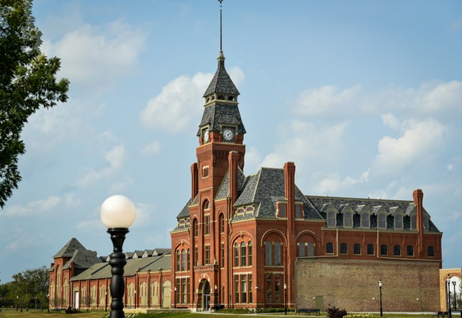 A red brick four story building with a clock tower from a distance with a round light pole in the foreground.