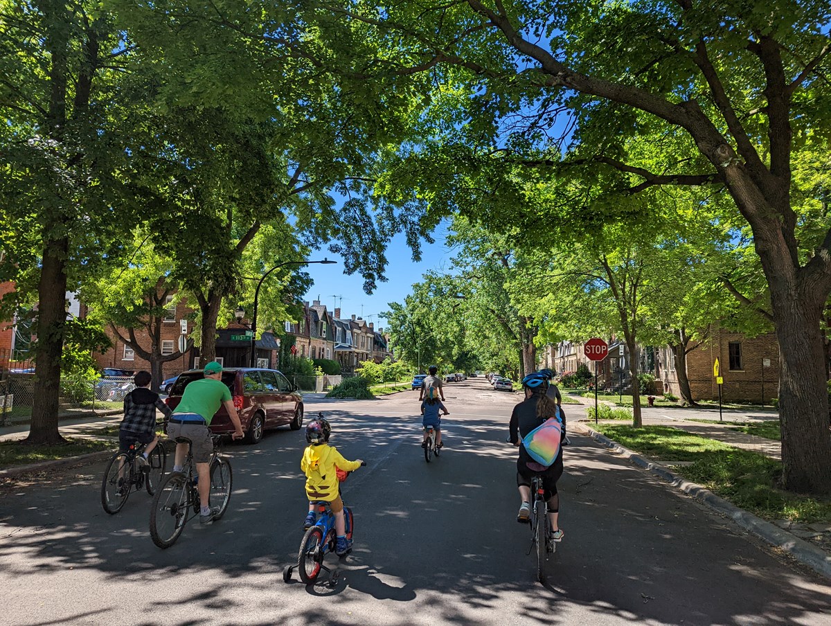 A ranger and visitors on bikes ride down a tree and historic rowhome lined street.