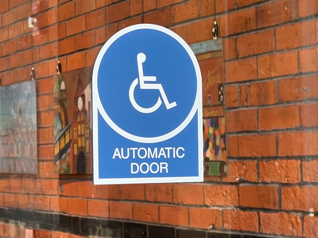 Photo of a blue Automatic Door Sign on a glass door. Behind the glass door are works of art along a brick wall.
